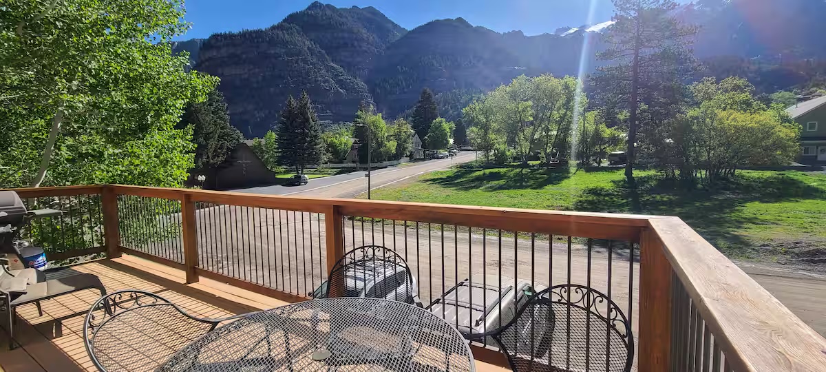 Deck with a patio table and chairs and a grill, looking out across the lawn and road to the mountains